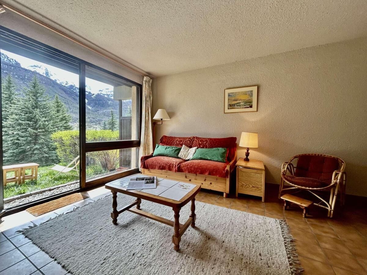 Ein gemütliches Wohnzimmer mit einem großen Fenster, einem Holztisch, einem roten Sofa, einem Korbstuhl und einem kleinen Tisch mit einer Lampe.
