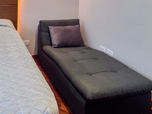 A single bed with a patterned bedspread, a dark sofa bed with a cushion, and a wall socket. - Casa com 4 Quartos - Alto Padrão (Carapicuíba)