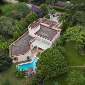 Uma casa grande, de vários andares, com piscina, cercada por vegetação exuberante.