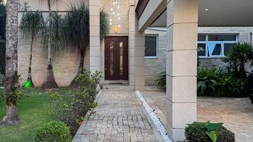 A modern building with a stone pathway leading to a wooden door, surrounded by greenery and a hanging light fixture.