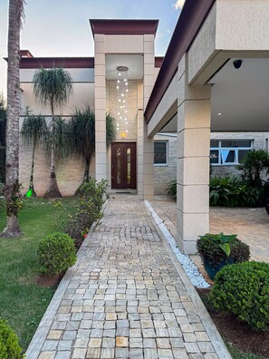 Un edificio moderno con un sendero de piedra que conduce a una puerta de madera, rodeado de vegetación y una lámpara colgante.