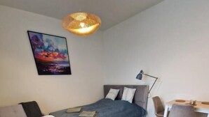 Un dormitorio con cama, mesita de noche, lámpara, silla, escritorio y un cuadro en la pared.