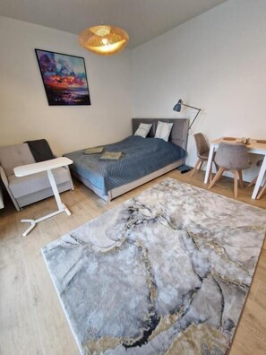 Ett sovrum med en säng, ett nattduksbord, en lampa, en stol, ett skrivbord och en tavla på väggen.