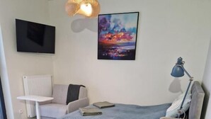 Un dormitorio con cama, mesita de noche, lámpara, silla y un cuadro en la pared.