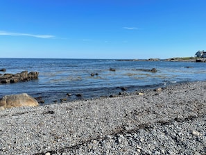 Eine felsige Küste mit klarem, blauem Wasser, vereinzelten Felsen und einem Gebäude in der Ferne.