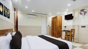 A hotel room with a bed, a desk, a chair, a TV, and a wall-mounted fan. - Hotel Elite INN (Dehradun)
