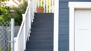 Un escalier avec des rampes blanches menant à une porte, le tout entouré d’un jardin où poussent différentes plantes et fleurs.