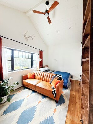 Een slaapkamer in loftstijl met een schuine plafond, een plafondventilator, een bed met een blauwe deken, een bruine leren bank en een groot raam met gordijnen.