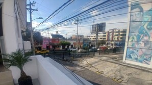 Un balcón con una maceta, vista de una calle con coches estacionados y un edificio con un mural.
