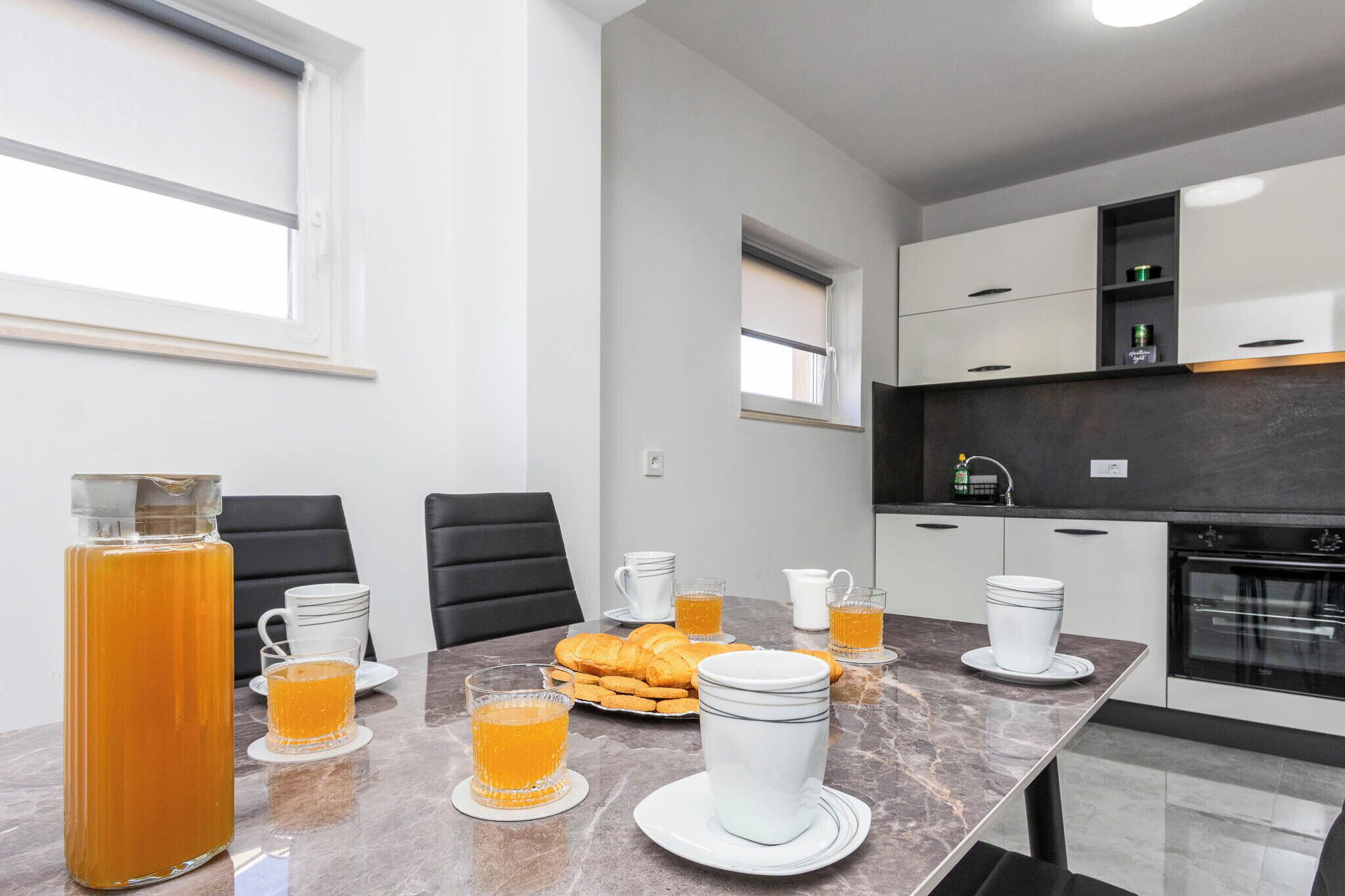 Une cuisine moderne avec des meubles blancs, un plan de travail en marbre et un coin repas où la table est mise pour le petit-déjeuner.