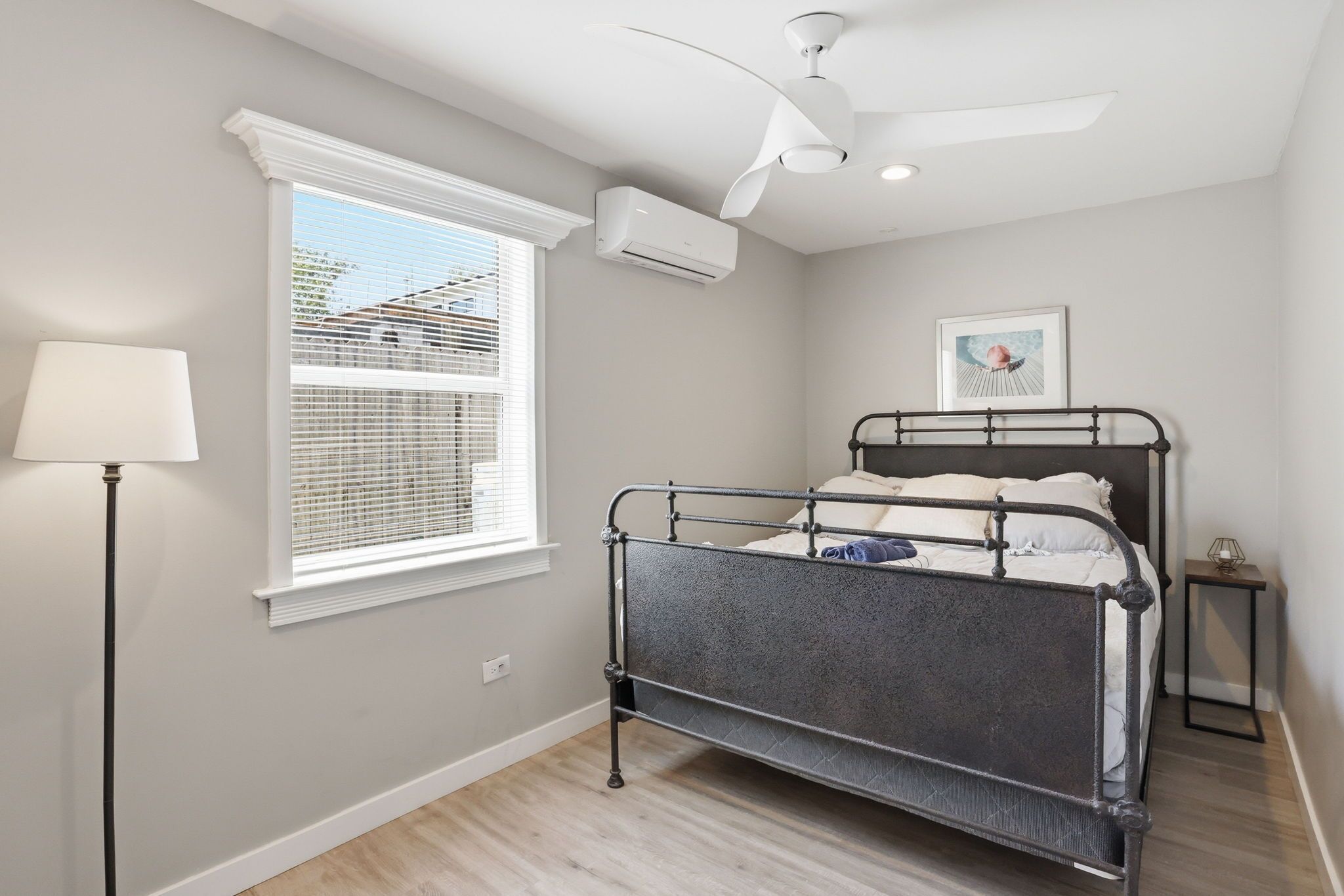 A bedroom with a metal bed frame, a ceiling fan, a window with blinds, and a bedside table with a lamp.