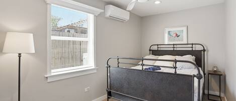 A bedroom with a metal bed frame, a ceiling fan, a window with blinds, and a bedside table with a lamp.