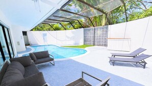 Un área de piscina exterior moderna con cerramiento de cristal, sillas de descanso y una parrilla incorporada.