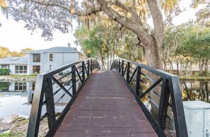 Eine Holzbrücke mit Metallgeländern führt zu einem Haus am Ufer eines ruhigen Gewässers.