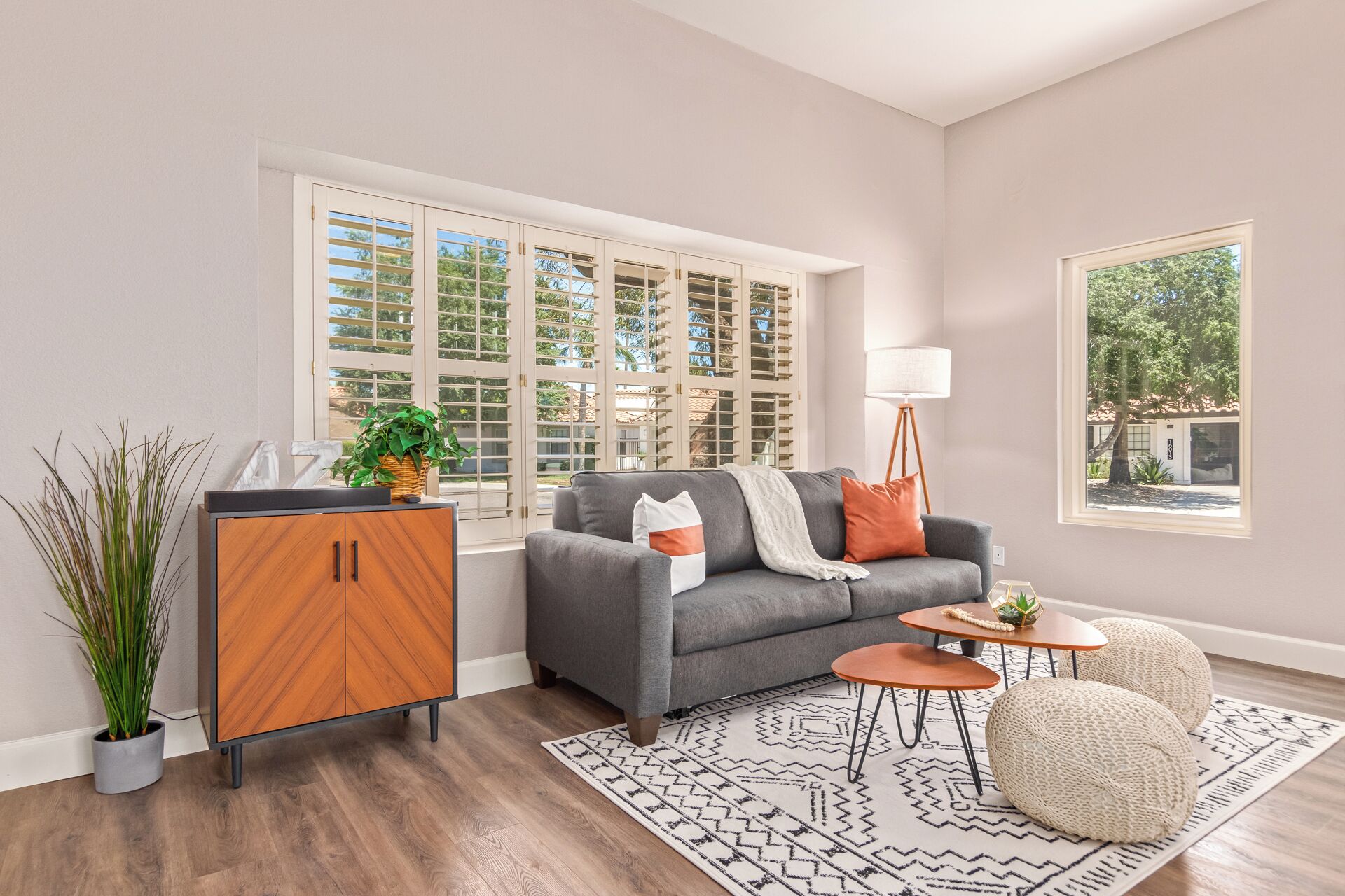 Ein modernes Wohnzimmer mit grauem Sofa, Holzschrank und großem Fenster mit Rollläden.