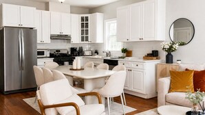 Een moderne keuken met witte kasten, een roestvrijstalen koelkast, een eethoek met een witte tafel en stoelen, en een zithoek met een bank en kussens.