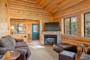 Ein Holzblockhaus-Innenraum mit Kamin aus Stein, einem darüber montierten Fernseher und einer gemütlichen Sitzecke mit Sofa und Hocker.