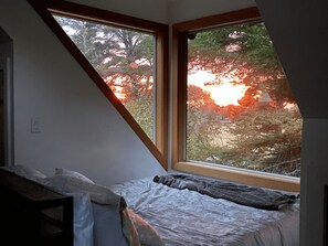 A bedroom with a sloped ceiling, a bed, and a window overlooking a forest.