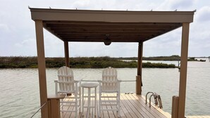Terraza de madera con dos sillas Adirondack y una mesita, con vista a un cuerpo de agua y humedales al fondo.