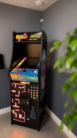 Una máquina arcade de Pac-Man en una habitación con paredes grises y una planta en primer plano.