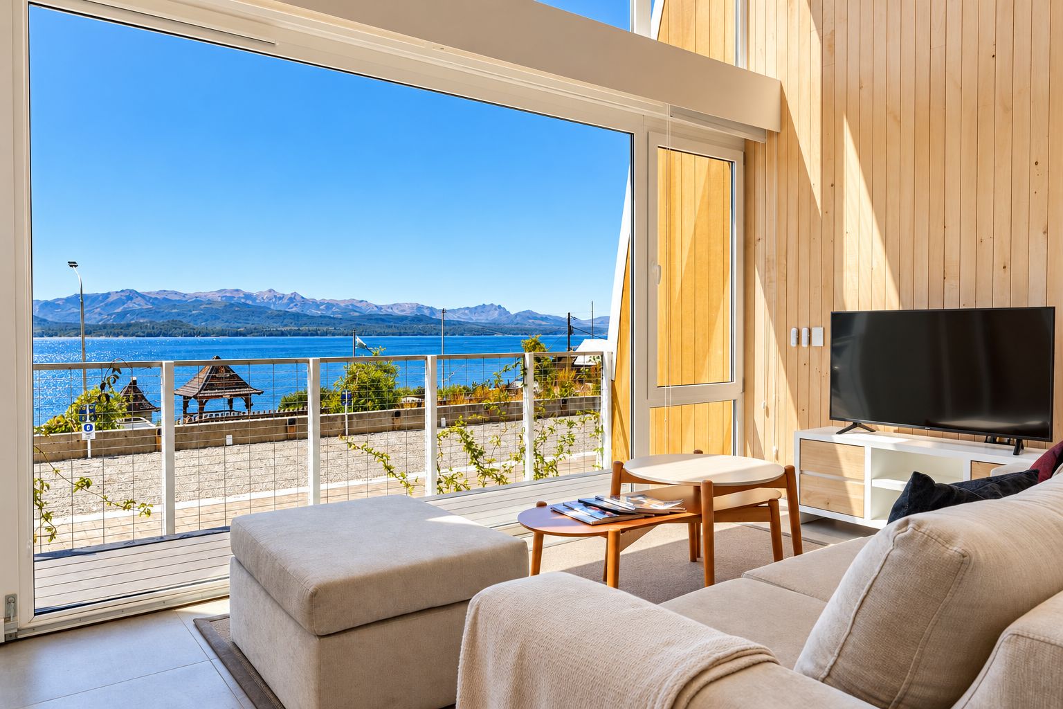 A modern living room with a large window overlooking a lake and mountains, a flat-screen TV, and a sofa.