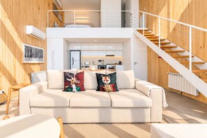 Ein modernes Wohnzimmer mit einer Couch, zwei Zierkissen, einer Holztreppe und einer Küche im Hintergrund.