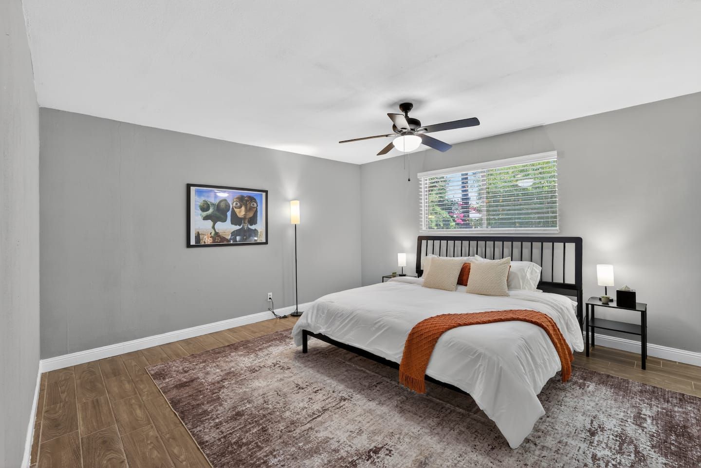 Une chambre à coucher avec un lit, un ventilateur de plafond, une fenêtre avec des stores, un tableau encadré au mur et une table de chevet avec une lampe.