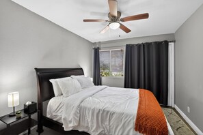 A hotel room with a bed, a nightstand, a lamp, a ceiling fan, and a window with curtains. - Heated Pool & Hot Tub | Old Town | Sleeps 10+ (Scottsdale)