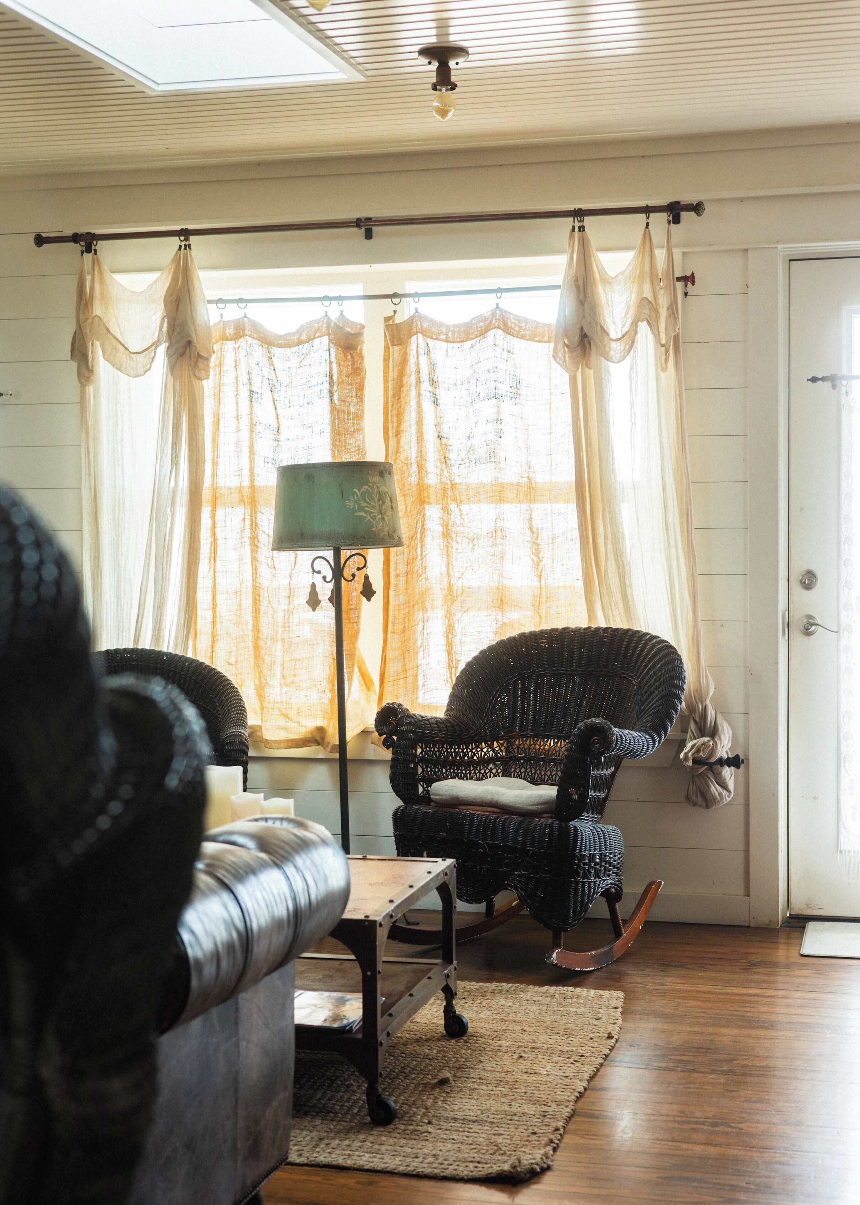 Una habitación con una silla de mimbre, una mesita pequeña, una lámpara de pie y una ventana con cortinas traslúcidas.