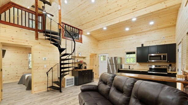 Interior de una cabaña con una escalera de caracol, una sala de estar con sofá de cuero, una cocina con electrodomésticos modernos y un ventanal con vistas a la vegetación.