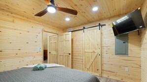 Habitación con paneles de madera, una cama, ventilador de techo y televisor montado.