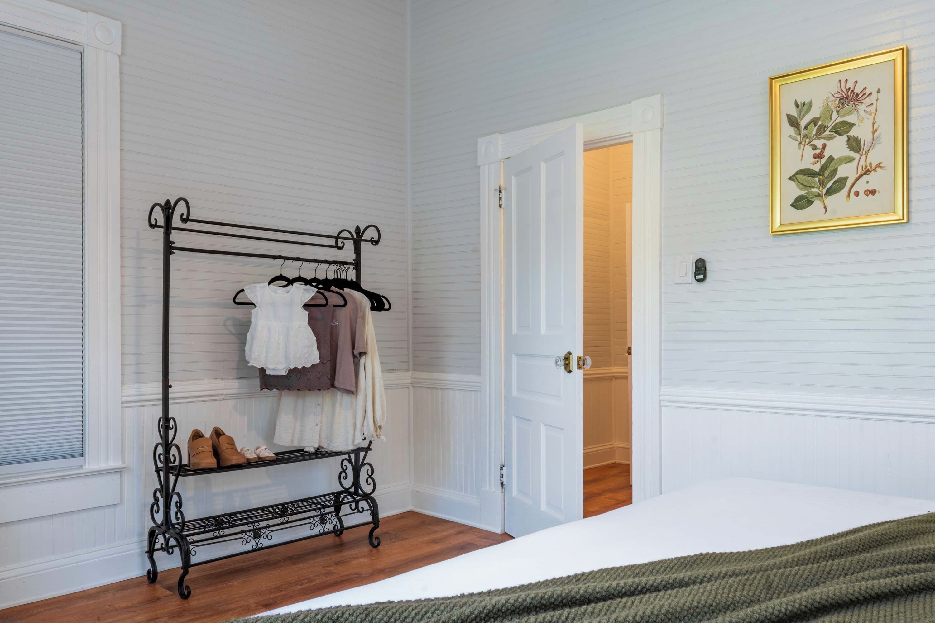 Une chambre avec un portant en métal où sont rangés des vêtements et des chaussures, un lit recouvert d’une couverture verte et une porte qui donne sur une autre pièce.