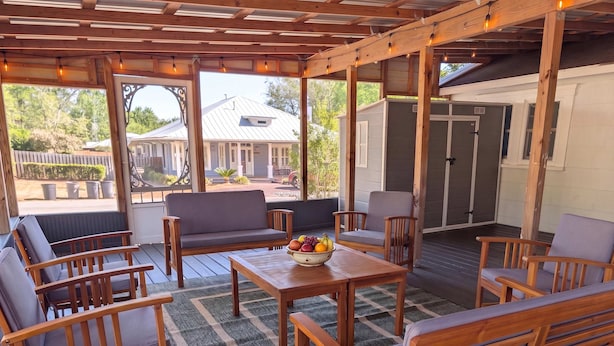 A covered patio with wooden furniture, a table with fruit, and a view of a house. - The Magnolia | 1900 Historic•Grand Piano•3 KingBRs (Alachua)