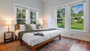A bedroom with a large bed, two bedside lamps, a nightstand, and a view of a green landscape through large windows. - The Magnolia | 1900 Historic•Grand Piano•3 KingBRs (Alachua)