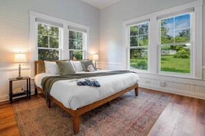 Een slaapkamer met een groot bed, twee bedlampen, een nachtkastje en uitzicht op een groen landschap door grote ramen.