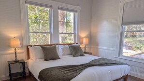A bedroom with a large bed, two bedside tables, two lamps, and a view of greenery through the windows. - The Magnolia | 1900 Historic•Grand Piano•3 KingBRs (Alachua)