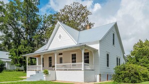 A two-story white house with a gabled roof, a front porch with a railing, and a small front yard. - The Magnolia | 1900 Historic•Grand Piano•3 KingBRs (Alachua)