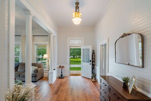 A hallway with a wooden floor, a sofa, a lamp, a table, and a mirror. - The Magnolia | 1900 Historic•Grand Piano•3 KingBRs (Alachua)