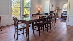 A dining area with a large wooden table and chairs, large windows with a view of trees, and a green pendant light. - The Magnolia | 1900 Historic•Grand Piano•3 KingBRs (Alachua)