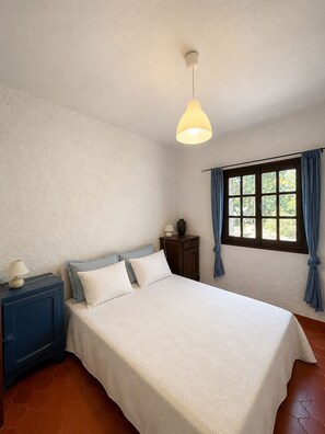 A bedroom with a bed, a nightstand, a lamp, a window with curtains, and a hanging light fixture. - Atypical Corsican villa with swimming pool and private beach access (Pianottoli-Caldarello)