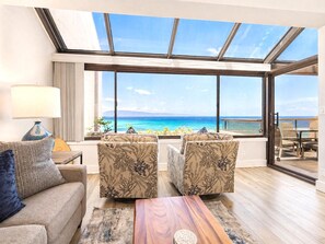 Ein Zimmer mit großem Fenster mit Blick auf einen Strand, eine Sitzecke, zwei Sessel, ein Couchtisch aus Holz und eine Lampe.
