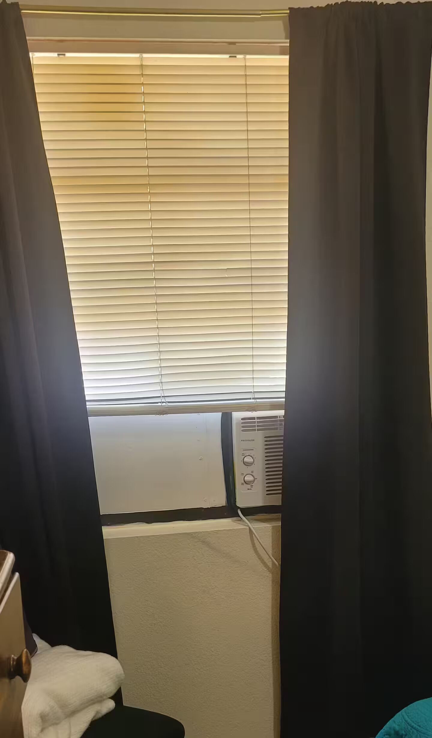 A room with a window with blinds, an air conditioning unit, and a towel on a chair.