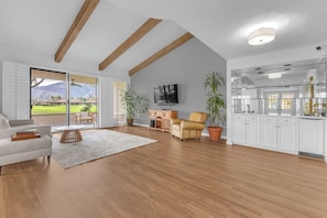 A spacious living room with a large window, a flat-screen TV, a sofa, a coffee table, and a kitchen area with white cabinets.