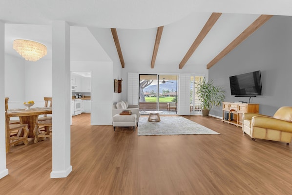 A spacious living area with a wooden floor, a sofa, a television, and a view of a garden through a sliding door.