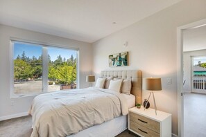 Une chambre avec une grande fenêtre, un lit, une table de chevet avec une lampe et un tableau au mur.