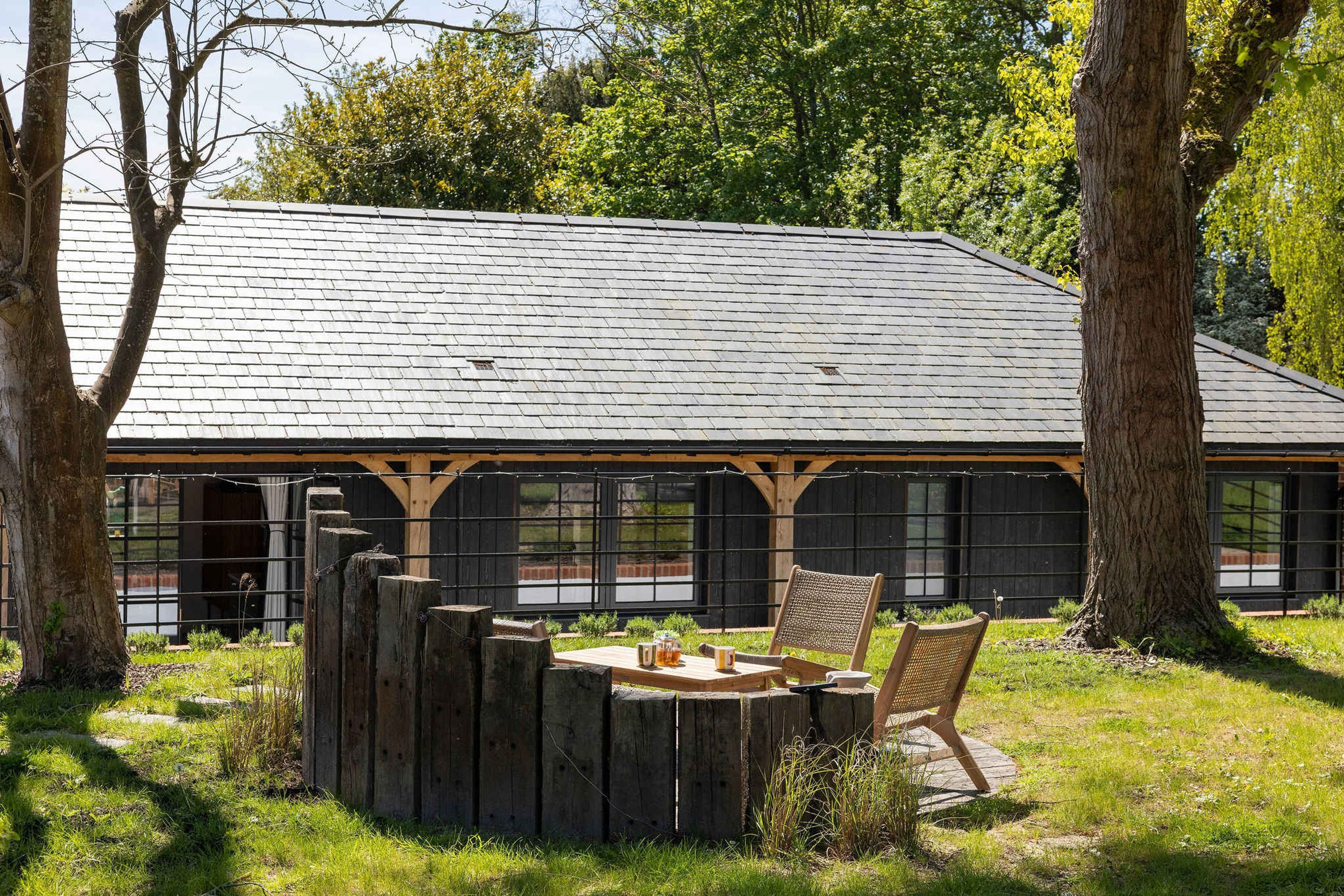 A rustic outdoor seating area with a wooden table and chairs, surrounded by trees and a building with a sloped roof.