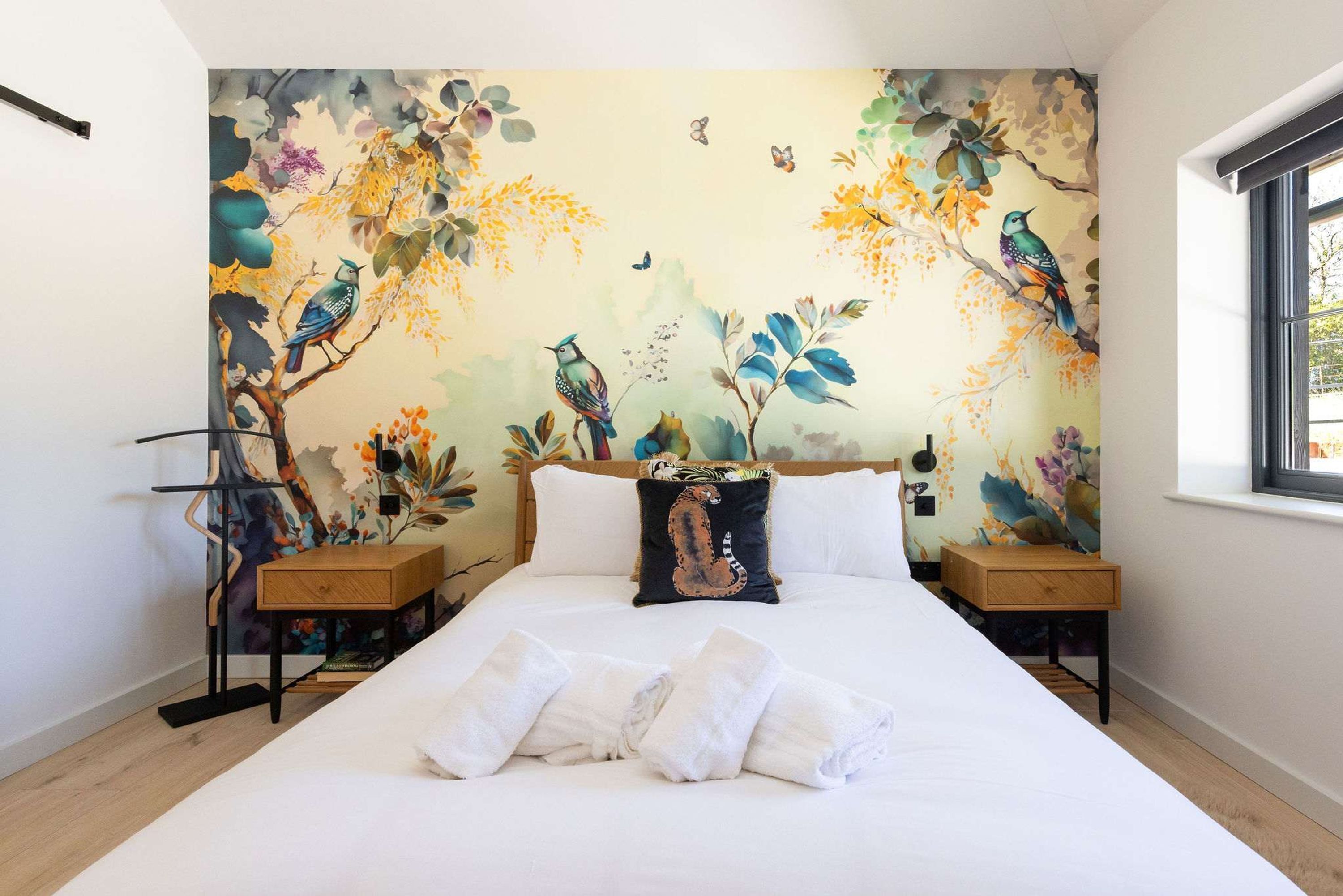 A bedroom with a large bed, two nightstands, a window, and a mural of birds and foliage on the wall.