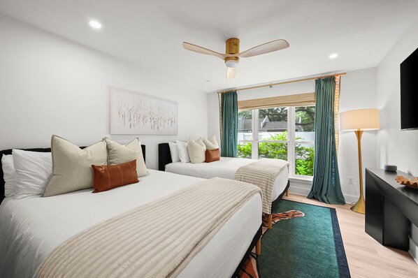 Habitación de hotel con dos camas, un ventilador de techo, un ventanal grande con cortinas, un televisor de pantalla plana y una lámpara de noche.