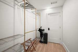 Una habitación con un tendedero metálico, una silla plegable de madera, una maleta negra y una puerta blanca.