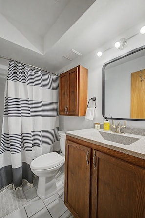 A bathroom with a striped shower curtain, a toilet, wooden cabinets, a sink with a mirror, and a towel rack. - Riverfront Ski & Fish Condo, Near BC & Vail (Edwards)